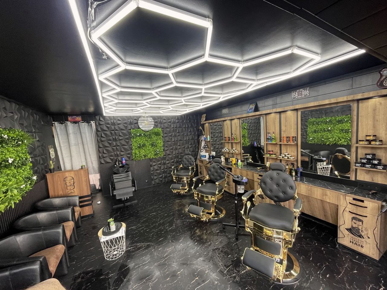 Le salon Sami’s Barber vu en entrant : plafond hexagonal lumineux, fauteuils de barbier noir et or, murs végétaux et sol en marbre noir